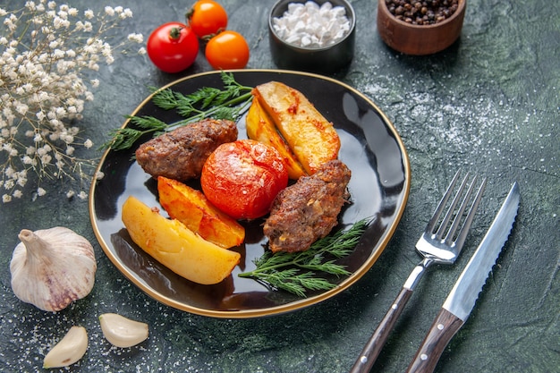 Vista frontale di deliziose cotolette di carne al forno con patate e pomodori su un set di posate piatto nero fiori bianchi spezie aglio su sfondo verde nero colori misti