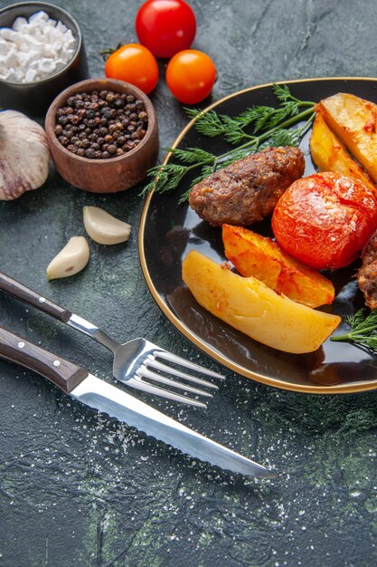 Vista frontale di deliziose cotolette di carne al forno con patate e pomodori su un set di posate piatto nero spezie garlics pomodori su sfondo verde nero colori misti