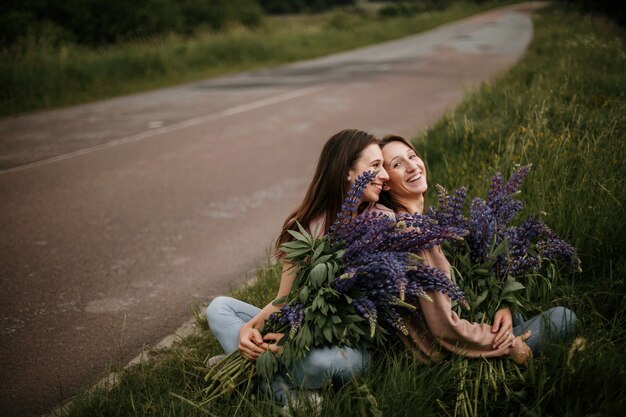 Vista frontale di due giovani ragazze brune sedute sull'erba con grandi mazzi freschi di lupini selvatici vicino alla strada e sinceramente sorridenti