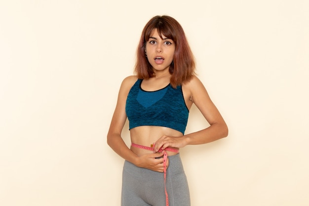 Vista frontale di giovane donna con corpo in forma in camicia blu che misura la sua vita sull'esercizio di allenamento atleta salute sport scrivania bianca luce