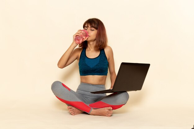 Vista frontale di giovane donna con corpo in forma in camicia blu utilizzando il suo laptop e acqua potabile sul muro bianco