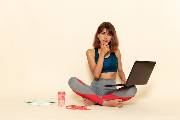 Vista frontale di giovane donna con corpo in forma in camicia blu utilizzando laptop sulla parete bianca leggera