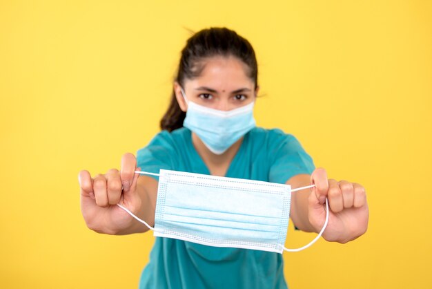 Vista frontale di giovane donna con maschera medica che tiene la maschera sulla parete isolata gialla