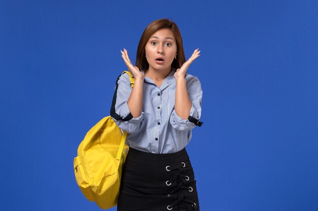 Vista frontale di giovane donna in camicia blu che porta zaino giallo in posa con espressione sorpresa sulla parete blu