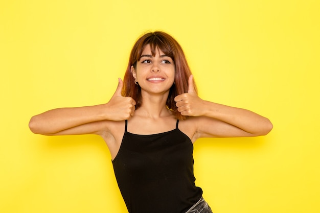 Vista frontale di giovane donna in camicia nera e jeans grigi sorridente sulla parete gialla