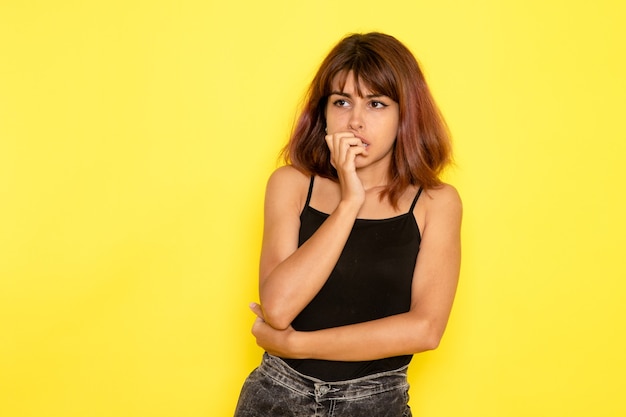 Vista frontale di giovane donna in camicia nera in posa con espressione confusa sulla parete gialla