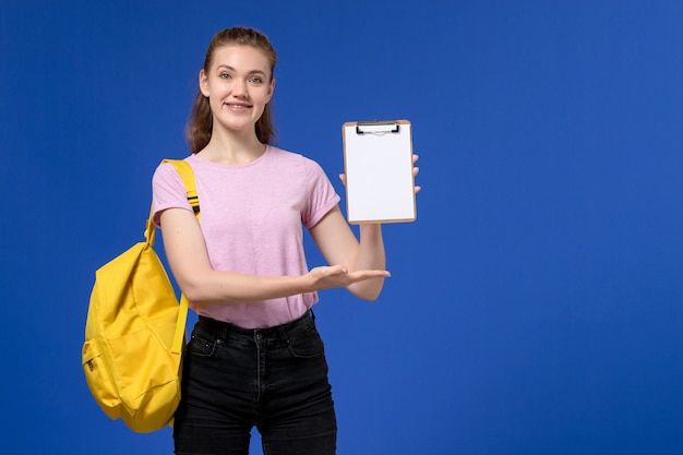 Vista frontale di giovane donna in maglietta rosa che indossa il blocco note sorridente della tenuta dello zaino giallo sulla parete blu