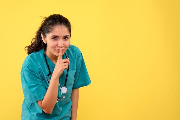 Vista frontale di giovane medico femminile che fa segno di sh sulla parete gialla