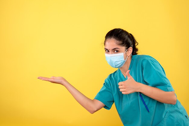 Vista frontale di giovane medico femminile con mascherina medica che fa il pollice sul segno sulla parete gialla