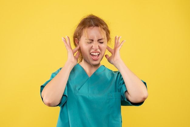 Vista frontale di giovane medico femminile in camicia medica che attacca le sue orecchie sulla parete gialla