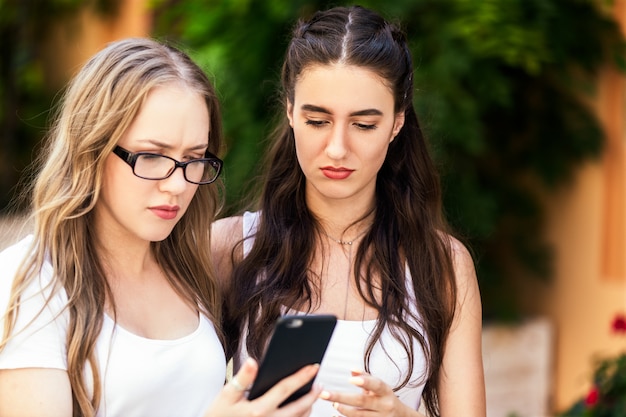 Vista frontale di giovani belle ragazze che guardano qualcosa al telefono con i fronti seri all'aperto al giorno caldo