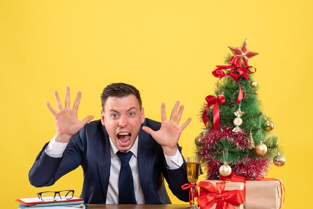 Vista frontale di gridare giovane uomo seduto al tavolo vicino albero di Natale e regali su giallo