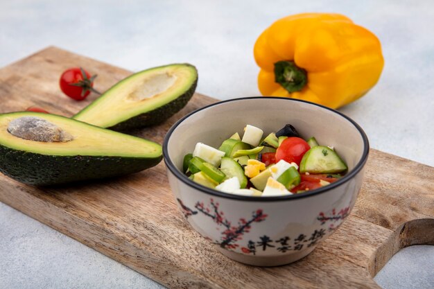 Vista frontale di insalata di verdure fresche con cetriolo pomodoro in una ciotola su una tavola di cucina in legno con fette di avocado e peperone giallo sulla superficie bianca