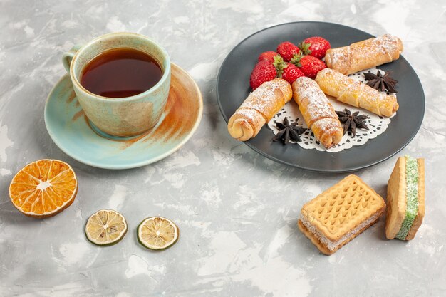 Vista frontale di panini in polvere di zucchero con fragole e tazza di tè sulla scrivania bianca