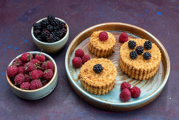 Vista frontale di piccole torte dolci e deliziose con frutti di bosco freschi sulla superficie scura