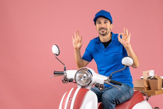 Vista frontale di un corriere felice sorridente che indossa un cappello seduto su uno scooter e fa il gesto degli occhiali su sfondo color pesca pastello