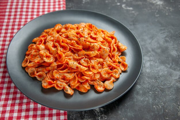 Vista frontale di un delizioso pasto di pasta su un piatto nero per cena su un asciugamano spogliato rosso su sfondo scuro