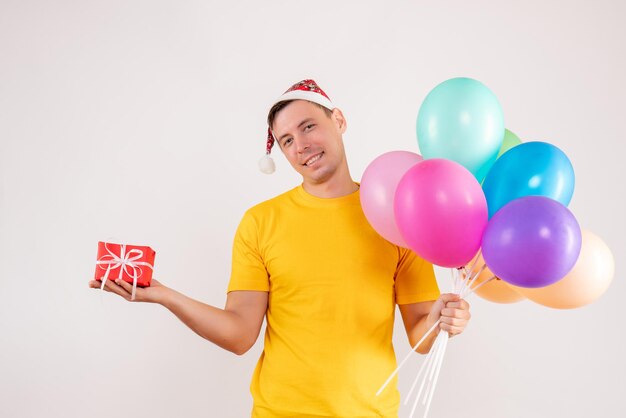 Vista frontale di un giovane che tiene in mano palloncini colorati e un piccolo regalo sul muro bianco