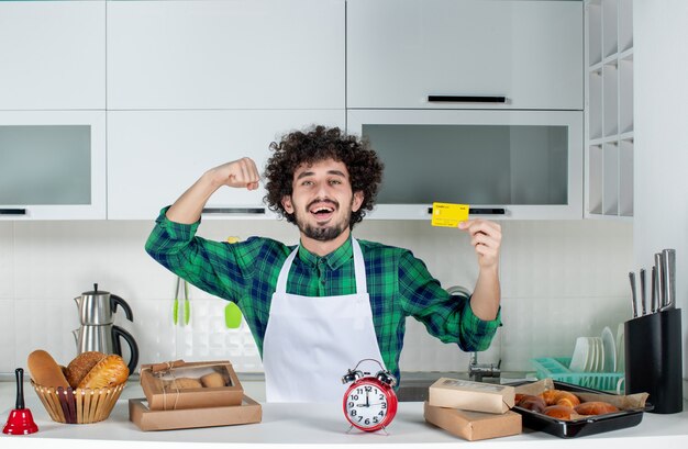 Vista frontale di un giovane fiducioso felice in piedi dietro il tavolo con vari pasticcini e che mostra la carta di credito nella cucina bianca