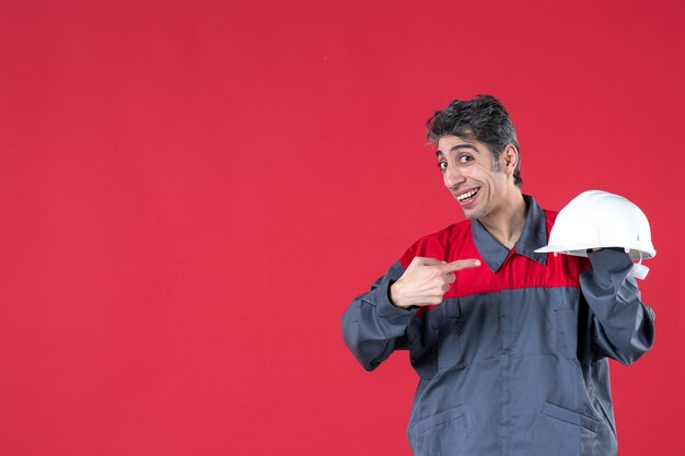 Vista frontale di un giovane lavoratore confuso in uniforme che punta elmetto sul muro rosso isolato