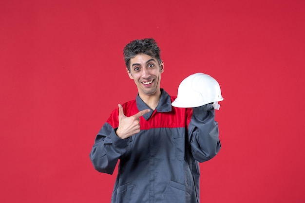 Vista frontale di un giovane lavoratore sorridente in uniforme e che punta elmetto sul muro rosso isolato