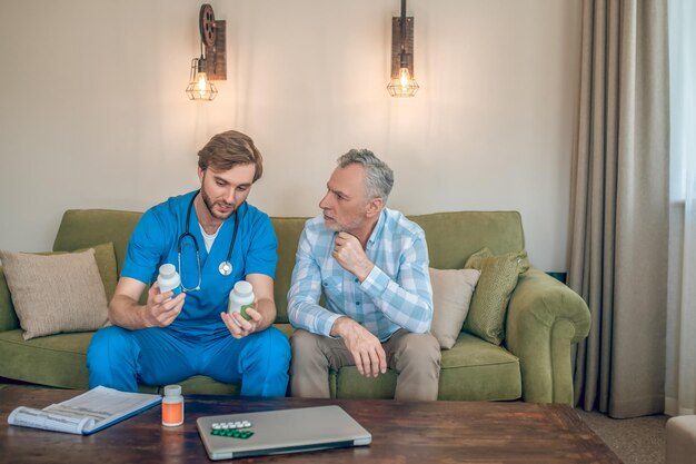 Vista frontale di un giovane medico con bottiglie di plastica di vitamine in mano che parla con un uomo maturo