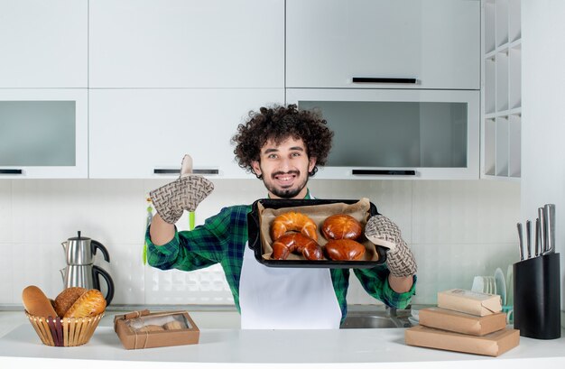 Vista frontale di un giovane ragazzo sorridente che indossa un supporto che mostra pasticceria appena sfornata che fa un gesto ok nella cucina bianca