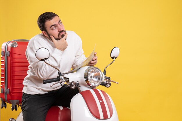 Vista frontale di un giovane uomo premuroso seduto su una moto con la valigia sopra che tiene una mappa su sfondo giallo isolato