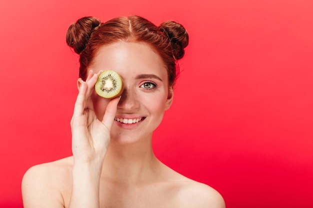 Vista frontale di una donna allo zenzero allegra con kiwi Foto in studio di una donna caucasica gioiosa con frutta esotica isolata su sfondo rosso