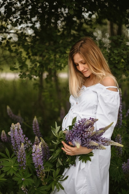 Vista frontale di una donna incinta calma vestita di abito bianco che tiene in mano un bouquet di lupini selvatici e guarda la pancia