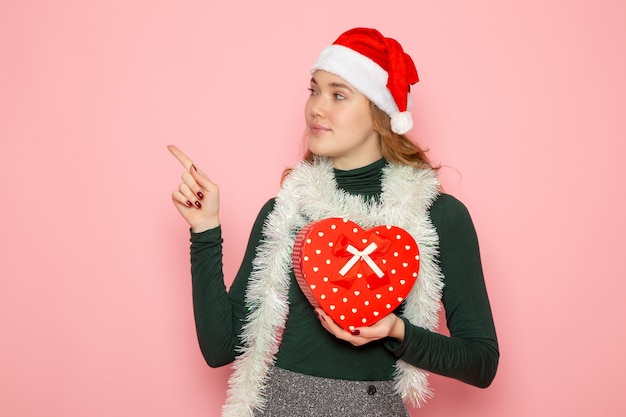 Vista frontale di una giovane donna con un berretto rosso che tiene un regalo a forma di cuore sul muro rosa