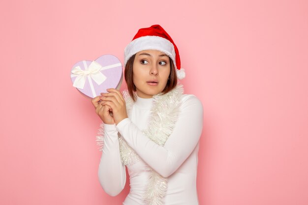 Vista frontale di una giovane donna con un berretto rosso che tiene un regalo a forma di cuore sul muro rosa