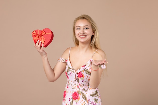 Vista frontale di una giovane donna in posa con un regalo a forma di cuore rosso sul muro marrone