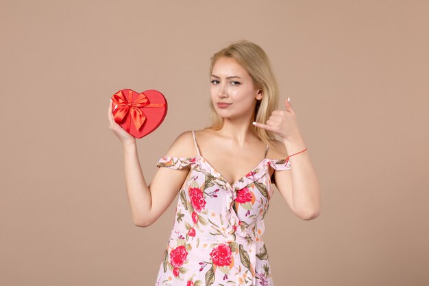 Vista frontale di una giovane donna in posa con un regalo a forma di cuore rosso sul muro marrone