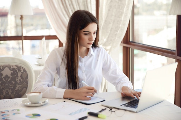 Vista frontale di una giovane imprenditrice bruna che sta lavorando sul computer portatile e scrivere qualcosa