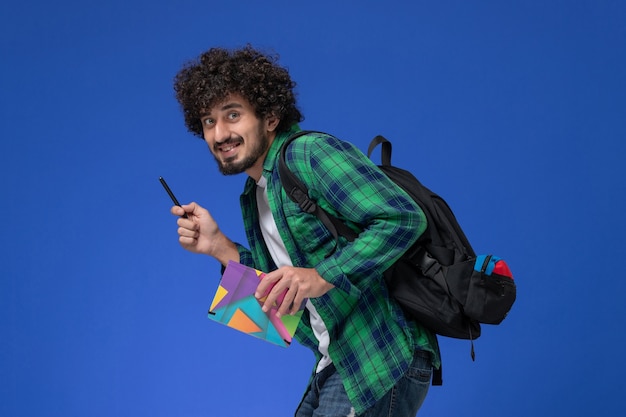 Vista frontale di uno studente maschio che indossa uno zaino nero che tiene il quaderno e la penna sulla parete azzurra