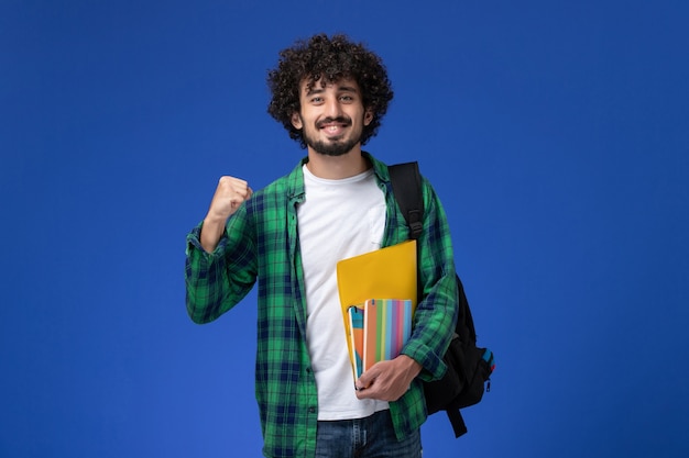 Vista frontale di uno studente maschio che indossa uno zaino nero con quaderni e file sulla parete blu