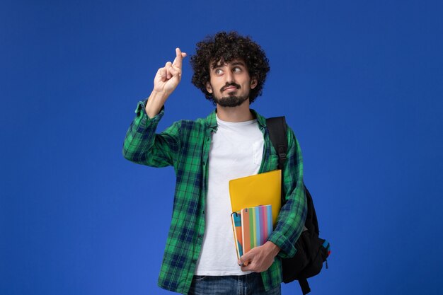 Vista frontale di uno studente maschio che indossa uno zaino nero con quaderni e file sulla parete blu