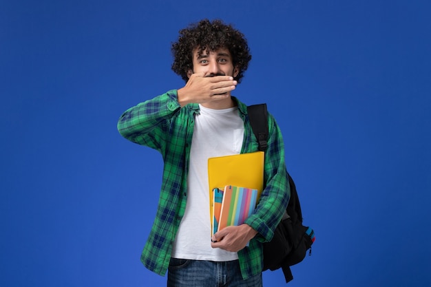 Vista frontale di uno studente maschio che indossa uno zaino nero con quaderni e file sulla parete blu