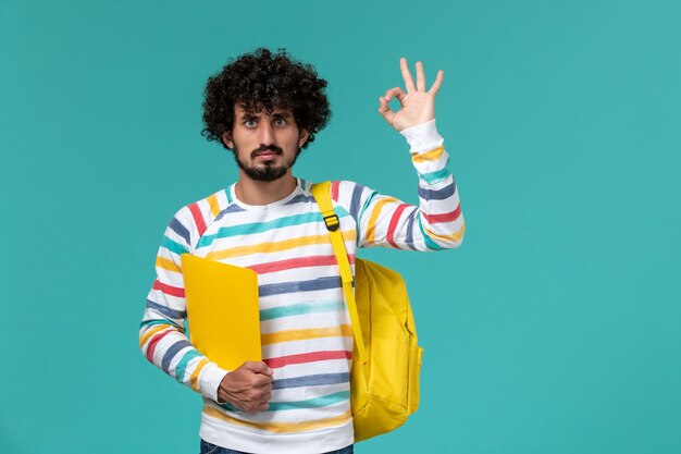 Vista frontale di uno studente maschio in camicia a righe che indossa lo zaino giallo che tiene i file sulla parete blu