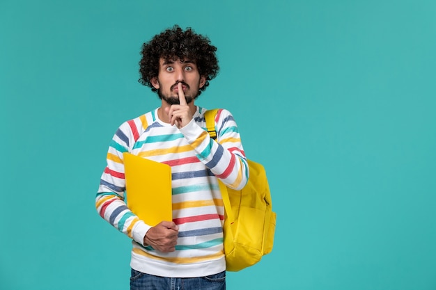 Vista frontale di uno studente maschio in camicia a righe che indossa lo zaino giallo che tiene i file sulla parete blu