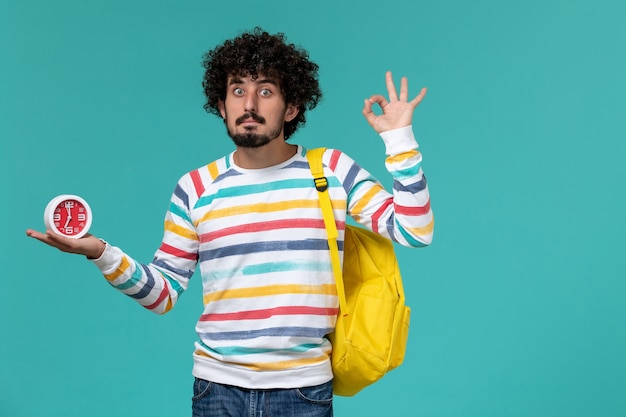 Vista frontale di uno studente maschio in camicia a righe che indossa uno zaino giallo che tiene gli orologi sulla parete blu