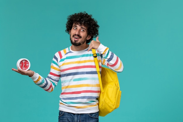 Vista frontale di uno studente maschio in camicia a righe che indossa uno zaino giallo che tiene gli orologi sulla parete blu