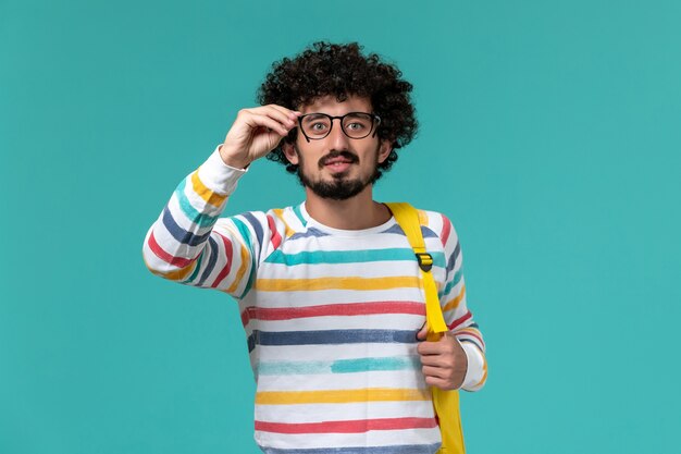 Vista frontale di uno studente maschio in camicia a righe che indossa uno zaino giallo che tiene i suoi occhiali da sole ottici sulla parete blu