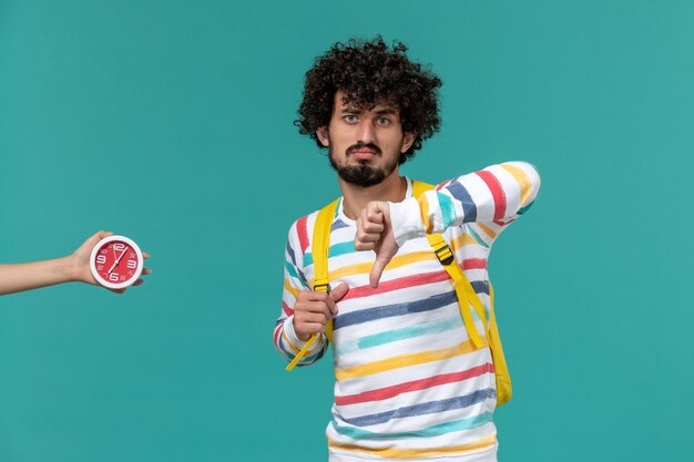 Vista frontale di uno studente maschio in camicia a righe che indossa uno zaino giallo in posa sulla parete blu chiaro