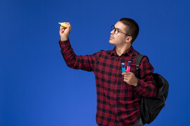 Vista frontale di uno studente maschio in camicia a scacchi rossa con zaino che tiene i pennarelli sulla parete blu chiaro