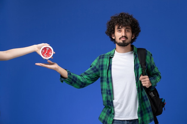 Vista frontale di uno studente maschio in camicia a scacchi verde che indossa uno zaino nero sulla parete blu chiaro