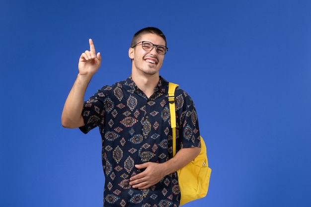 Vista frontale di uno studente maschio in camicia di cotone scuro che indossa uno zaino giallo in posa e ridendo sulla parete azzurra