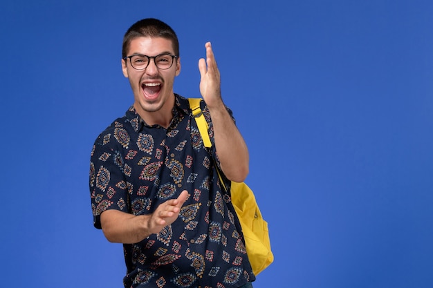 Vista frontale di uno studente maschio in camicia scura che indossa uno zaino giallo che ride ad alta voce sul muro azzurro