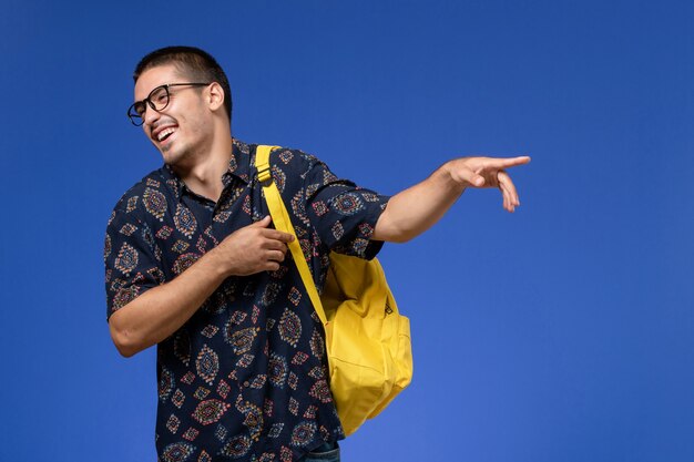 Vista frontale di uno studente maschio in camicia scura che indossa uno zaino giallo e ridendo sulla parete blu
